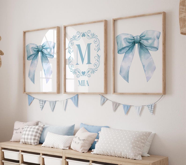May include: Three framed prints with a blue watercolor bow and the letter M in a decorative frame with the name Mia below. The prints are hanging on a white wall above a wooden bench with pillows.
