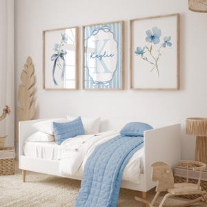 May include: Three framed wall art prints with watercolor floral designs and a personalized name. The prints feature light blue flowers and ribbons, with a wooden frame. The prints are displayed above a white bed with blue pillows and a blue quilt.