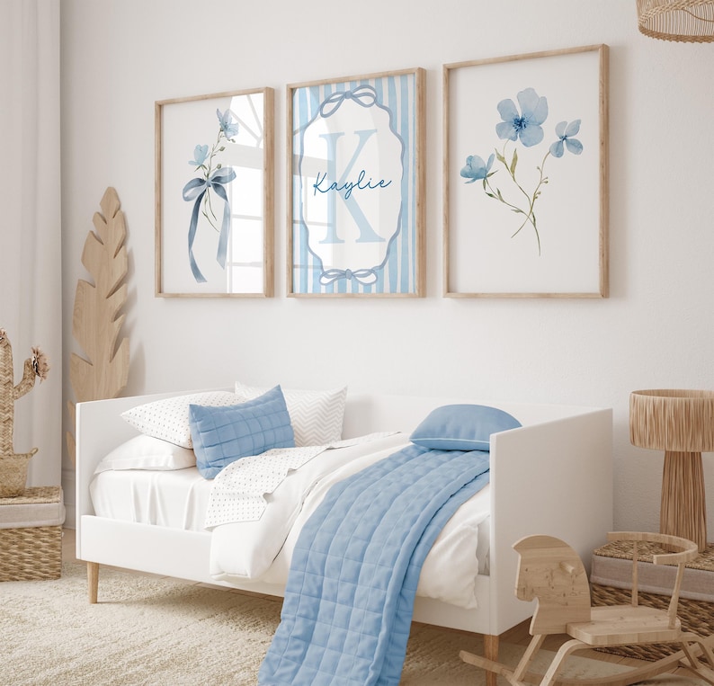 May include: Three framed wall art prints with watercolor floral designs and a personalized name. The prints feature light blue flowers and ribbons, with a wooden frame. The prints are displayed above a white bed with blue pillows and a blue quilt.