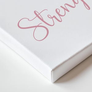 May include: White canvas art print with the word "Strength" in a cursive, dusty rose font. The canvas is stretched over a wooden frame, with the edges visible. The artwork is set against a white background.