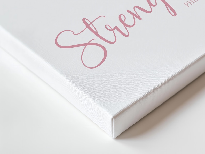 May include: White canvas art print with the word "Strength" in a cursive, dusty rose font. The canvas is stretched over a wooden frame, with the edges visible. The artwork is set against a white background.