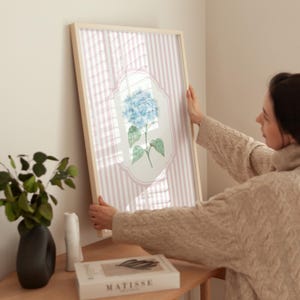 May include: A framed floral print featuring a blue hydrangea blossom. The artwork is set against a pink and white striped background, encased in a light wood frame. The print is being held up against a wall.
