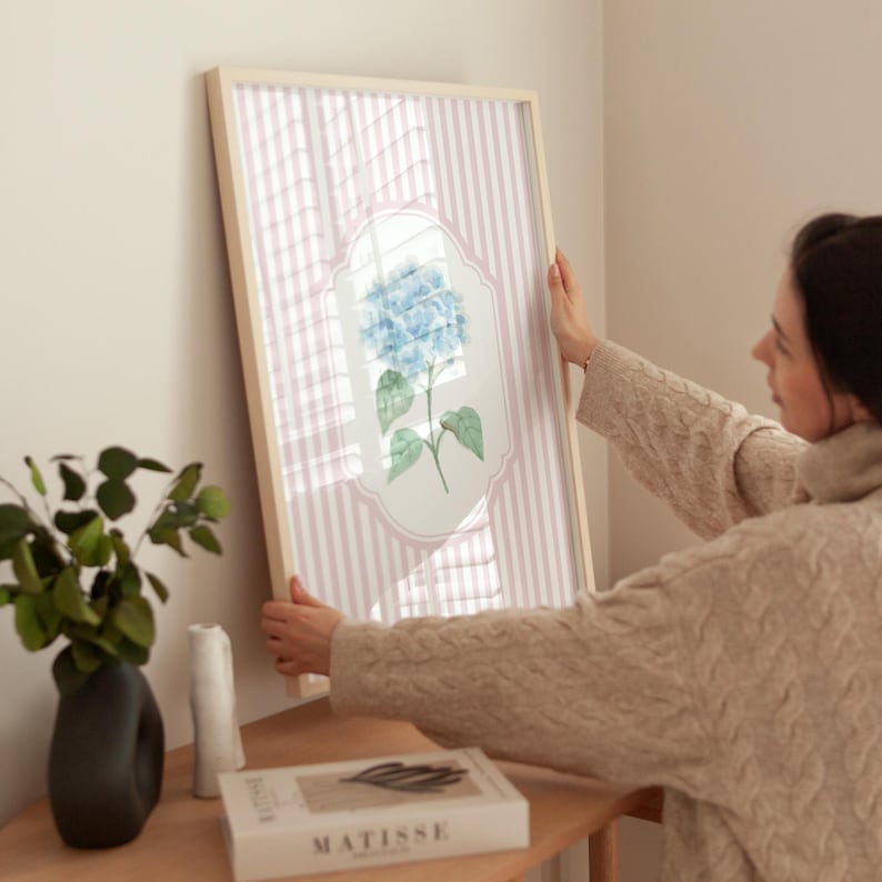 May include: A framed floral print featuring a blue hydrangea blossom. The artwork is set against a pink and white striped background, encased in a light wood frame. The print is being held up against a wall.