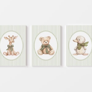 May include: Three framed art prints with watercolour illustrations of a giraffe, teddy bear, and duck. Each animal has a green bow and is set against a striped background. The frames are rectangular with an oval border.