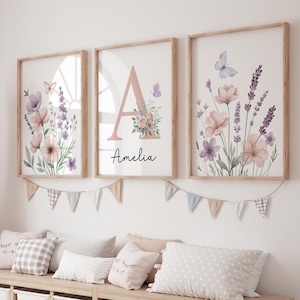 May include: Three framed floral art prints with watercolor illustrations of flowers and butterflies in soft pink, purple, and green hues. One print features the letter "A" and the name "Amelia". The frames are light wood.