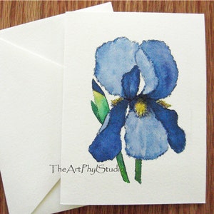 May include: A watercolor painting of a blue iris flower with a yellow center. The flower is painted in a realistic style and has a delicate, flowing appearance. The painting is signed "The Art Phyl Studio" in the bottom right corner.