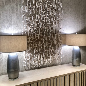 May include: Two lamps with beige lampshades and silver bases sit on a wooden table with a light wood finish. A white, string-like wall hanging with multiple loops hangs on the wall behind the table.