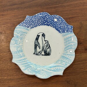 Op de afbeelding: Een wit keramiek bord met een blauwe en witte gepatroneerde rand. Het midden van het bord heeft een zwart-witte illustratie van twee pinguïns.