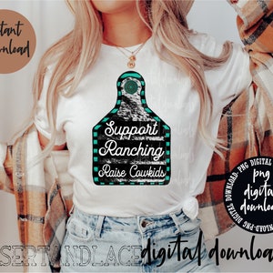 May include: A white t-shirt featuring a black and white cow tag design. The tag has teal blue trim and the text "Support Ranching Raise Cowkids".