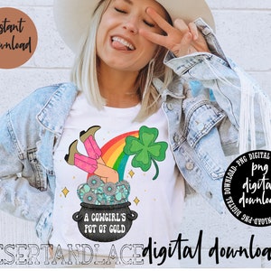 Pode incluir: Camiseta branca com um gráfico de arco-íris, um trevo de quatro folhas e um pote de ouro com o texto "A Cowgirl's Pot of Gold". A camisa também tem o texto "Desert and Lace" e "digital downloads".