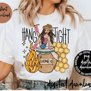 May include: A white t-shirt with a graphic design of a cartoon woman with a cowboy hat and boots, sitting on a honey jar with a flame behind her. The text "HANG TIGHT" is above the graphic and "HONEY" is on the jar.