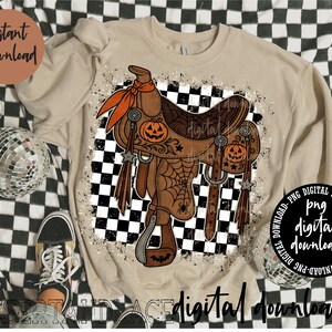May include: A beige crewneck sweatshirt featuring a graphic print of a brown western saddle decorated with orange jack-o'-lanterns, spider webs, and stars. The design is set against a black and white chequered background with a distressed paint splatter effect.