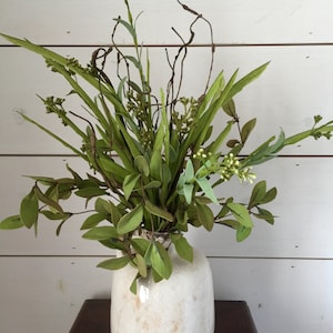 May include: A white ceramic vase with a textured surface holds a bouquet of green leafy branches and twigs. The vase has a rounded shape and a brown base.