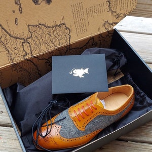 May include: A pair of brown leather shoes with a blue and white patterned toe. The shoes are in a black box with a black dust bag and a card with a fish logo.
