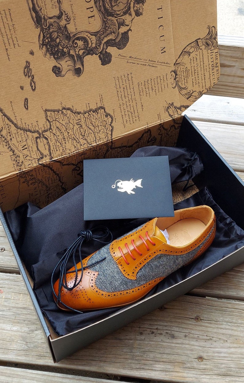 May include: A pair of brown leather shoes with a blue and white patterned toe. The shoes are in a black box with a black dust bag and a card with a fish logo.