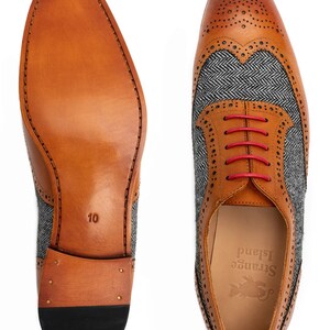 May include: A pair of brown leather shoes with a grey tweed pattern on the sides. The shoes have red laces and a black sole. The shoes are size 10 and have the brand name "Strange Island" on the inside.