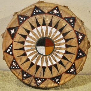 May include: A round hand drum with a tan rawhide surface featuring a central circle divided into four quadrants of black, red, white, and yellow. The drum head is decorated with a radiating pattern of white and black triangles and star shapes.