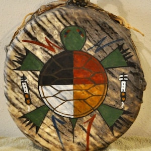May include: A round hand drum with a textured, mottled brown and white hide surface. The center features a painted turtle with a shell divided into four quadrants of black, red, white, and orange. Green limbs and red and blue lightning symbols surround the turtle.