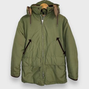 Puede incluir: Parka de invierno verde oliva con capucha, cierre de cremallera y dos bolsillos delanteros. La parka presenta detalles marrones en el forro de la capucha, los bolsillos y los puños. Una prenda de abrigo cálida y clásica.