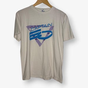 Puede incluir: Una camiseta blanca con el texto "CAPTAIN EO" en azul y morado. El texto está dentro de un triángulo morado. La camiseta está colgada en una percha de madera.