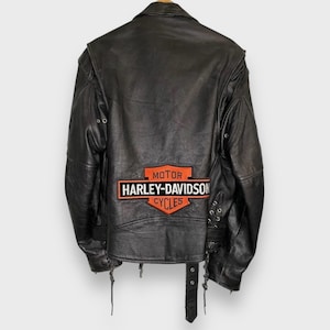 May include: Black leather motorcycle jacket with the Harley-Davidson Cycles logo patch on the back. The jacket has a notched collar, shoulder epaulets, and a belt at the waist. The jacket is a classic style.