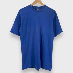 May include: A solid blue t-shirt with short sleeves hangs from a wooden hanger. The shirt has a crew neck and a simple, classic design. The fabric appears to be soft and slightly worn.