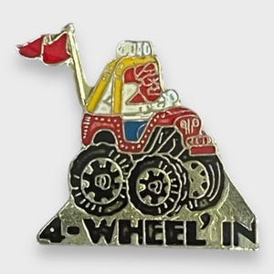 Puede incluir: Un colorido pin de esmalte con un vehículo todoterreno de estilo caricatura con grandes neumáticos negros. El vehículo es rojo, amarillo y azul, con una bandera roja. El pin dice "4-WHEEL'IN" en letras negras.