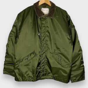 Puede incluir: Chaqueta militar verde oliva con cuello de pana marrón. La chaqueta tiene dos bolsillos delanteros, cierre de botón y palanca, y forro acolchado. Cuelga de una percha de madera.