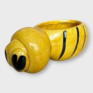 May include: A yellow ceramic bee-shaped planter with black stripes and heart-shaped eyes. The planter has a glossy finish and a hollowed-out opening. The bee design is a charming decorative item.