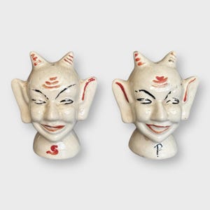 May include: A pair of vintage devil-shaped salt and pepper shakers. The ceramic shakers are off-white with red and black accents. Each has a smiling face, horns, and large ears. The salt shaker is marked with an "S", and the pepper shaker with a "P".