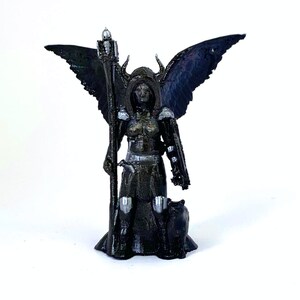 Puede incluir: Una figura miniatura negra y plateada de una figura femenina con alas, sosteniendo un bastón y de pie con un gato negro a sus pies.