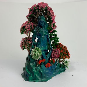 May include: A blue figurine of a woman standing in a forest setting. The figurine is surrounded by pink, red, and green trees. The base of the figurine is a green, rocky surface.