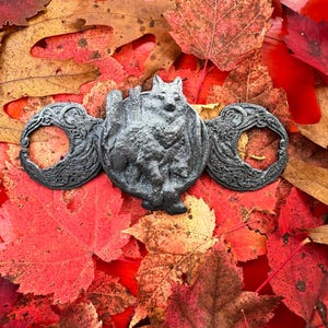 May include: A black metal pendant featuring a wolf standing on a crescent moon with a tree behind it. The pendant is surrounded by two more crescent moons with intricate designs. The pendant is lying on a bed of red and orange autumn leaves.