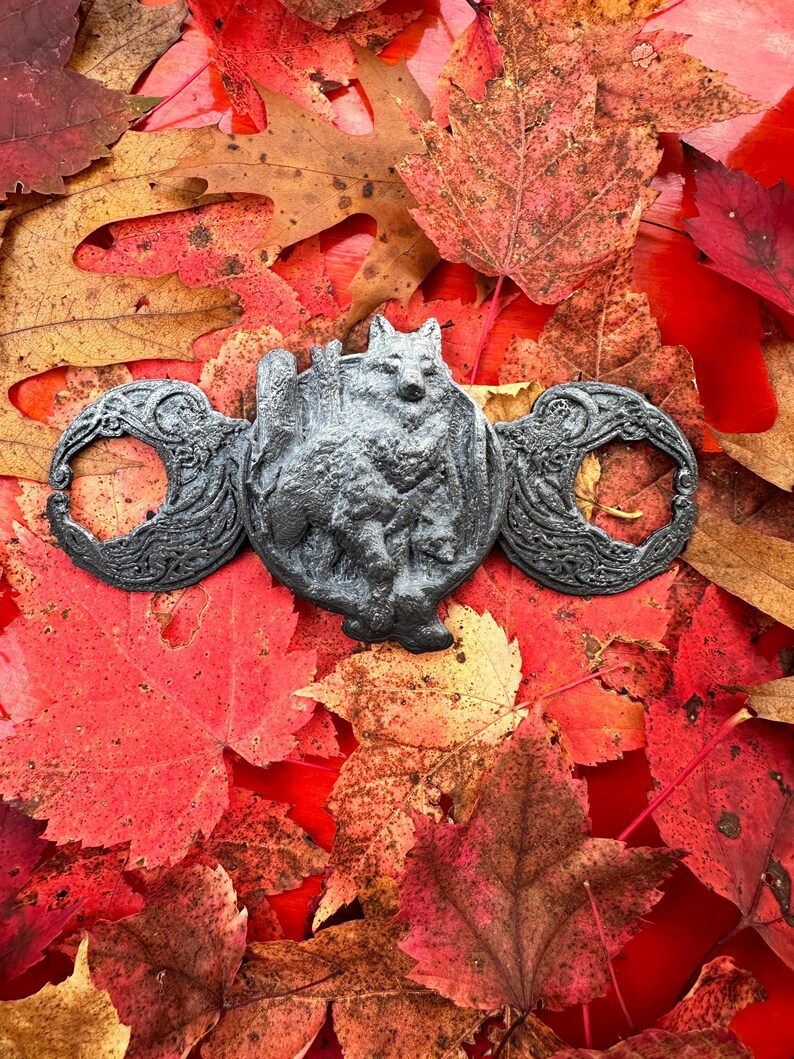 May include: A black metal pendant featuring a wolf standing on a crescent moon with a tree behind it. The pendant is surrounded by two more crescent moons with intricate designs. The pendant is lying on a bed of red and orange autumn leaves.