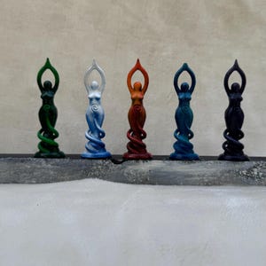 May include: Five small, colourful statues of women in a standing pose with their arms raised. The statues are painted in shades of green, white, orange, blue, and black.