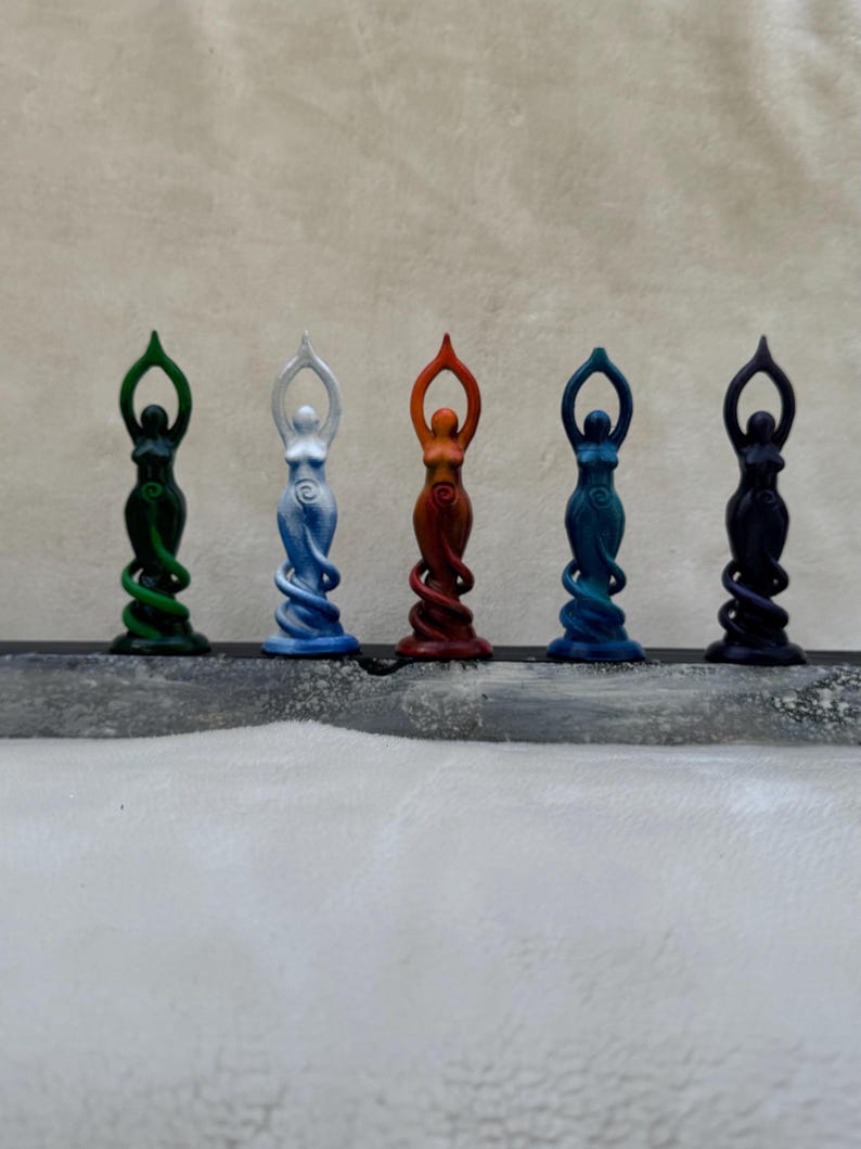 May include: Five small, colourful statues of women in a standing pose with their arms raised. The statues are painted in shades of green, white, orange, blue, and black.