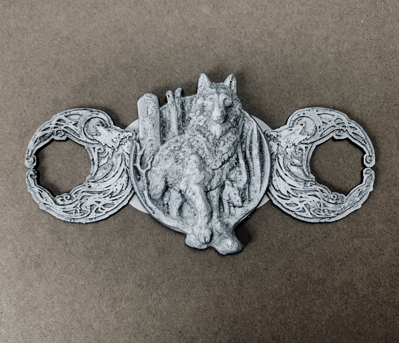 May include: A silver-toned metal pendant featuring a wolf design with intricate details. The wolf is standing on a crescent moon with a floral pattern. The pendant is attached to a chain.