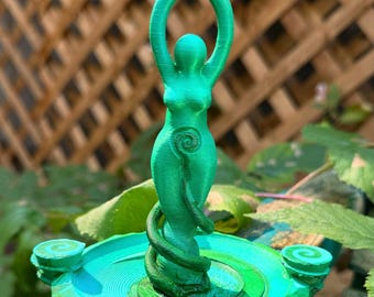 May include: A bright green 3D-printed figurine of a stylized human figure with arms raised above the head, standing on a circular base. The figure features a spiral design on the torso and a vine-like element wrapping around the lower body.