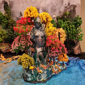 May include: A 3D printed figurine of a woman standing in a forest setting. The woman is made of a dark gray material and is surrounded by trees with red, yellow, and green leaves. The base of the figurine is covered in small, colorful flowers.