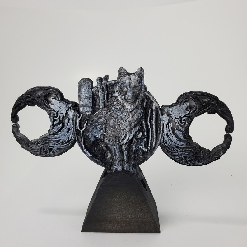 May include: A black and silver 3D printed sculpture of a wolf standing on a crescent moon. The wolf is surrounded by a decorative design of leaves and vines. The sculpture is mounted on a black wooden base.