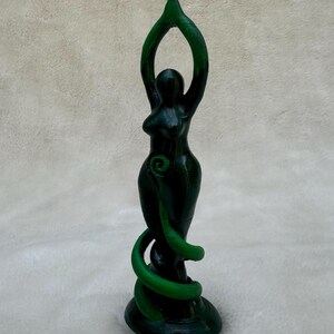 May include: Green goddess statue with arms raised in a pose of worship. The statue is made of a dark green material and has a swirling design.