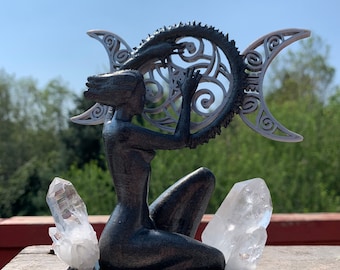May include: A black and silver 3D printed sculpture of a woman holding a snake in a circle with a triple moon symbol. The sculpture is sitting on a black base with two clear quartz crystals.