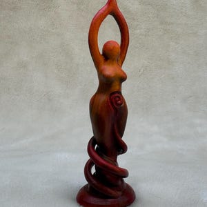 May include: A red and orange wooden sculpture of a woman with her arms raised above her head. The sculpture is standing on a base with a spiral design.