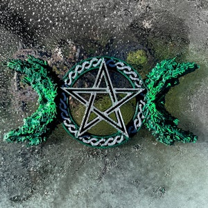 May include: A silver pentacle with a green triple moon symbol on a gray background.