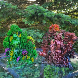 May include: Two 3D printed sculptures of figures standing in a forest setting. The figure on the left is green and surrounded by green and yellow foliage. The figure on the right is red and surrounded by red and brown foliage.