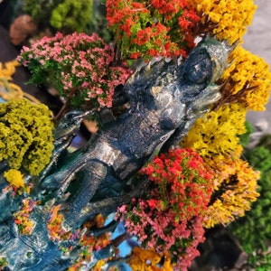 May include: A blue and silver figurine of a woman standing in a forest setting. The figurine is surrounded by miniature trees with pink, red, and yellow flowers.