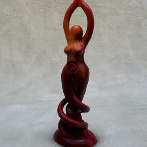 May include: A wooden sculpture of a woman with her arms raised, painted in shades of red and orange. The sculpture has a spiral design on the woman's abdomen.