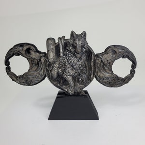 May include: A silver-colored sculpture of a wolf standing on a black base. The wolf is surrounded by a decorative design of swirling lines and shapes. The sculpture is a unique and eye-catching piece of art.