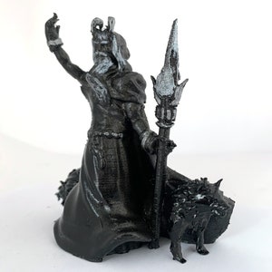 Puede incluir: Una miniatura impresa en 3D en negro y plata de una figura femenina que lleva una t&uacute;nica larga y sostiene un bast&oacute;n. La figura est&aacute; de pie sobre una base con un lobo a sus pies.