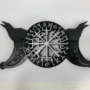 Puede incluir: Decoración de pared negra y plateada con una brújula Vegvisir rodeada de dos medias lunas con cabezas de cuervo. La brújula está rodeada de runas.